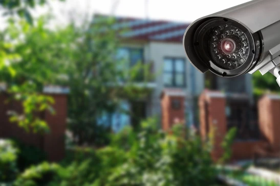 Caméra de surveillance extérieure installée dans un environnement résidentiel avec végétation luxuriante et bâtiments en arrière-plan